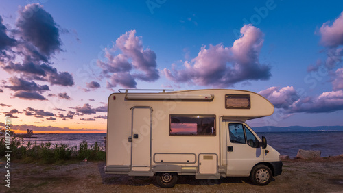 Camper van parked by the sea at sunset, under a colorful sky with dramatic clouds. Peaceful evening atmosphere, perfect for travel, adventure, freedom, and outdoor lifestyle themes