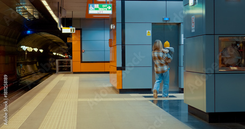 Young mother gently holding infant while walking across subway platform, moving toward elevator in urban transit setting
