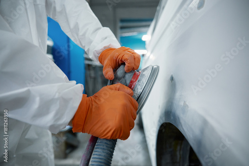 An auto body technician sands a car panel in a protective suit and orange gloves