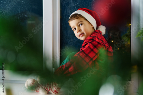 A little boy in a red shirt and hat sits on a window sill, waiting for Santa Claus in a decorated room. Concept for celebrating New Year and Christmas.