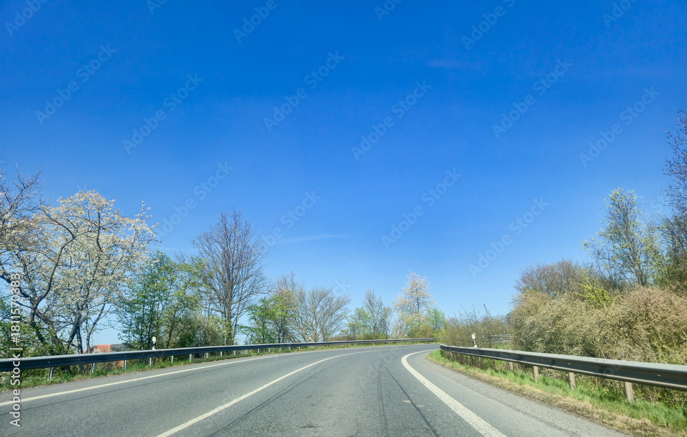 Fototapeta premium Curved road surrounded by trees under a clear blue sky, showcasing the beauty of nature in spring, ideal for travel and adventure themes.