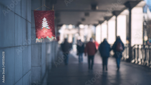 Fototapeta Naklejka Na Ścianę i Meble -  A small, illuminated Christmas tree banner hangs on a wall, while blurred people walk along a covered urban pathway, signaling holiday spirit.
