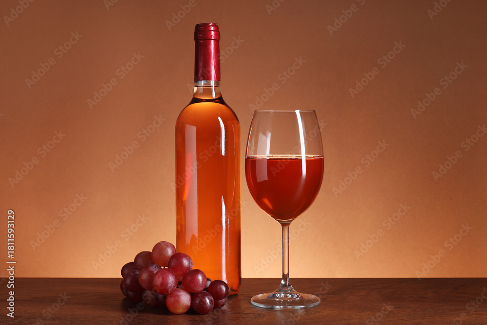 Fototapeta premium Rose wine in glass, bottle and grapes on wooden table against brown background