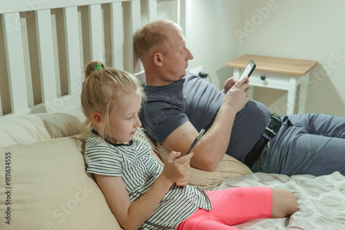 Man and girl child relaxing on bed using separate smartphones. Digital addiction in family life. Screen time and modern technology.