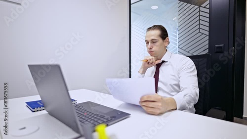 A focused businessman in a crisp white shirt and tie carefully reviews documents at his office desk, contemplating the details. He holds a pencil, deep in thought, with a laptop nearby.