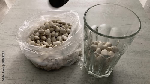 Kitchen Task: A close-up of a person using a tablespoon to transfer dry white beans from a faceted glass cup into a clear plastic bag for storage or portioning.