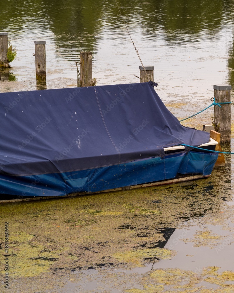 Fototapeta premium Small moored boat covered with tarpaulin in green water lake