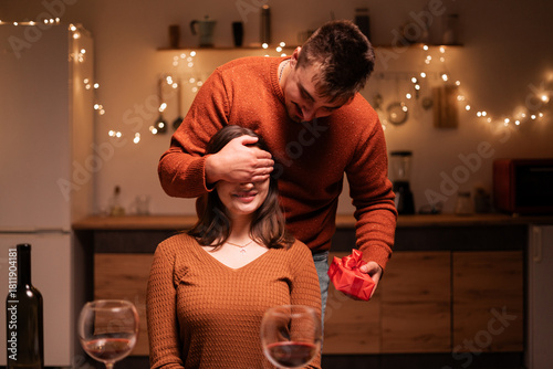 Surprise Gift for lover woman. Romantic man covering girlfriends eyes giving present box as greeting with birthday, anniversary or valentines day in the kitchen on home interior