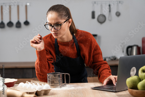 Positive woman learns a new recipe online using laptop at a kitchen looking ingredients on table