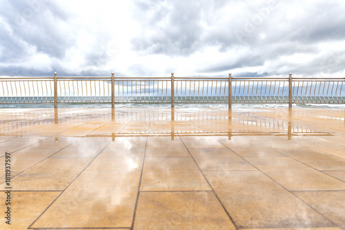 Fototapeta Naklejka Na Ścianę i Meble -  A view of the Baltic Sea from the embankment. The gray cloudy sky and metal fence are reflected in rain puddles on the yellow concrete pavement. Background. Landscape.