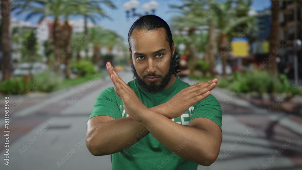Fototapeta premium Man with crossed arms forming an x gesture, hands and forearms visible, wearing a green shirt while standing on a sunny street lined with palm trees; rejection protest.