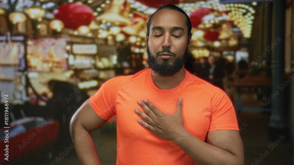 Fototapeta premium Man touching his hand to chest in street market with orange tshirt, lanterns and festive lights; serenity gratitude.
