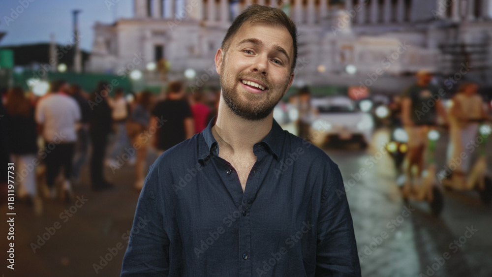 Fototapeta premium Man with visible face and trimmed beard tilts head and smiles while standing amidst a crowded street with blurred traffic at dusk; contemplation.