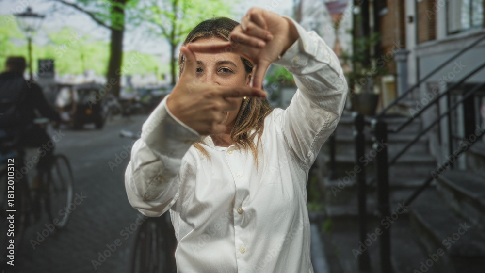 Obraz premium Young woman in white shirt framing face with hands on city street near stairs and lamppost; creative playful confidence.