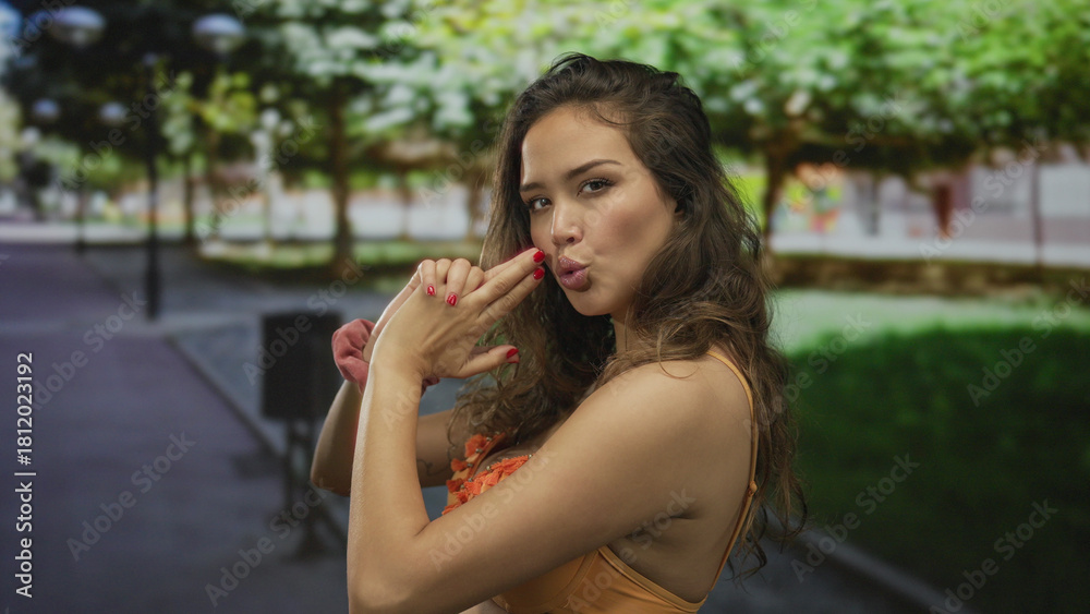 Naklejka premium Young hispanic woman in orange bikini pointing finger like pistol at camera in a park; playful confidence.