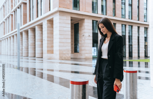 Wallpaper Mural Casual elegance by a modern building, a woman in a black outfit strolls, holding a red phone, amidst sleek urban lines and polished cityscape. Torontodigital.ca