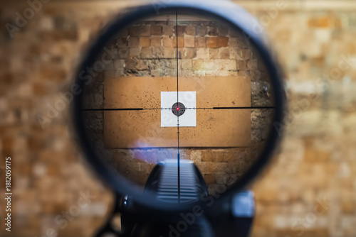 View through a sniper rifle scope aiming at a paper target with bullet holes in an indoor shooting range