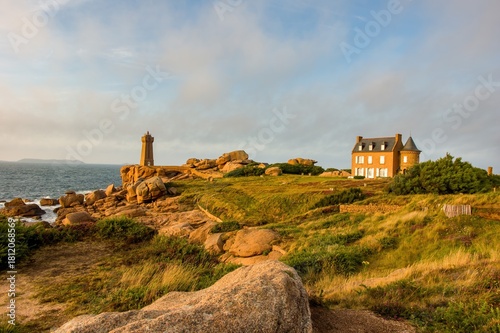 A lighthouse and historic stone house overlook a rocky coastline at sunset. Golden light illuminates the rugged landscape, ocean waves, and dramatic clouds, creating a serene coastal atmosphere.