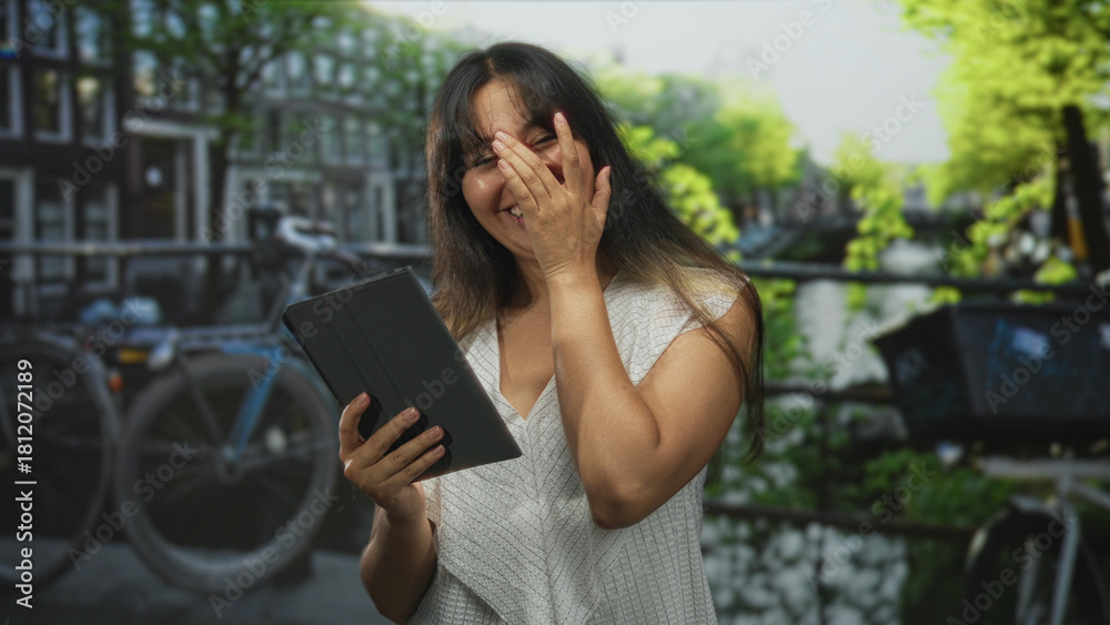 Fototapeta premium Woman holding tablet and hand covering face while smiling on a street by a canal in amsterdam outdoors; joy connection.