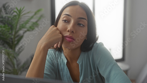 Young woman resting hand on chin at home interior near window and laptop leaning on desk; pensive reflection. Free Download Young woman resting hand on chin at home interior near window and laptop leaning on desk; pensive reflection. - GFXLoad