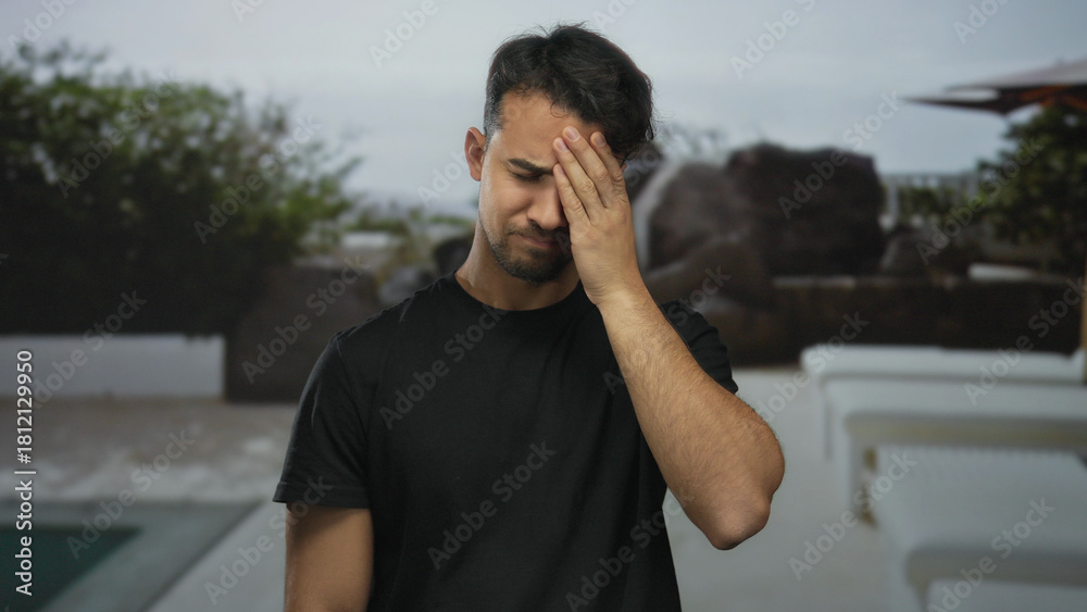 Fototapeta premium Young hispanic man at a resort pool outdoor appearing stressed or in discomfort, set against the tranquil backdrop of a hotel poolside scene.