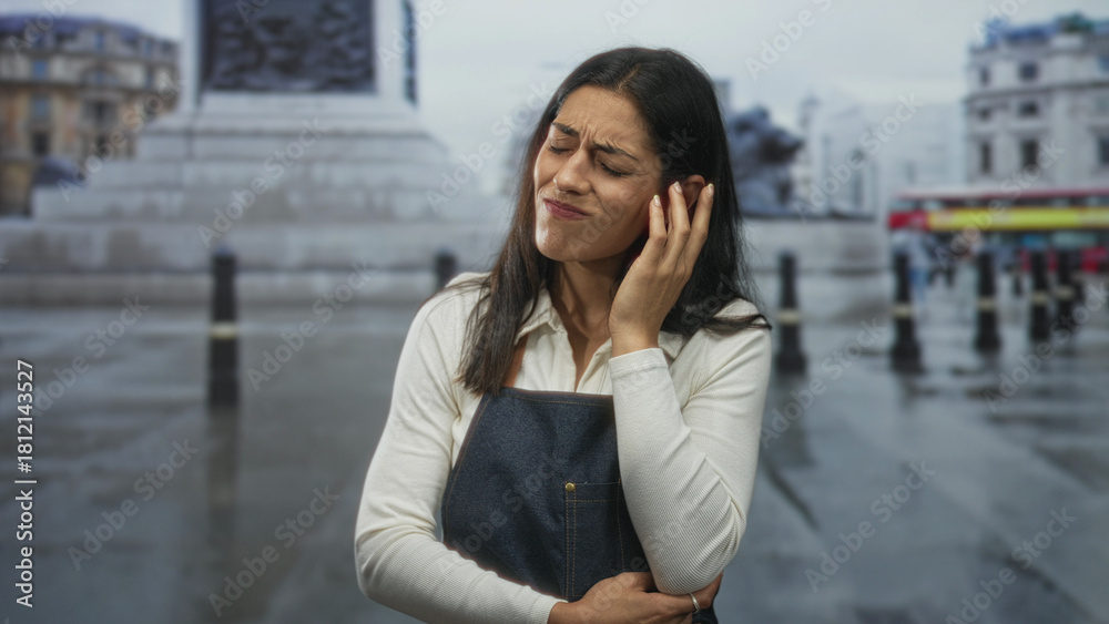 Fototapeta premium Woman holding hand to ear on wet street beside stone fountain and bollards, wearing denim apron, grimacing in pain; confusion.