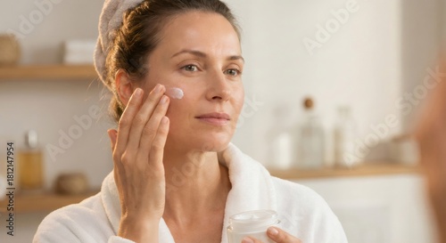 Woman in a bathrobe applying moisturizer to her face, reflecting her commitment to self care and beauty in a bathroom setting