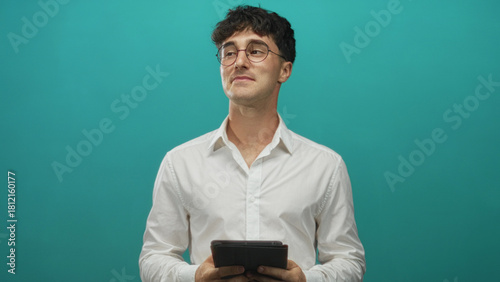 Man holding tablet and looking up with a slight smile in a teal studio wearing a white shirt and round glasses; thoughtful curiosity ambition.