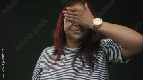 Woman wearing striped shirt covers eyes with hand in dark green studio setting; secrecy concealment embarrassment.