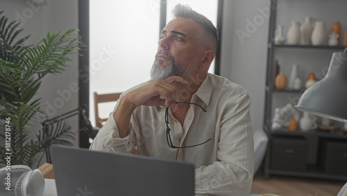Vászonkép Man holding glasses with hand on chin at desk in home office, gazing upward in a pensive pose beside laptop and lamp; thoughtful reflection