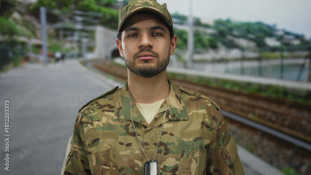 Fototapeta premium Young hispanic soldier stands at a railway station platform with camouflage uniform and visible dog tags on his chest; determination.