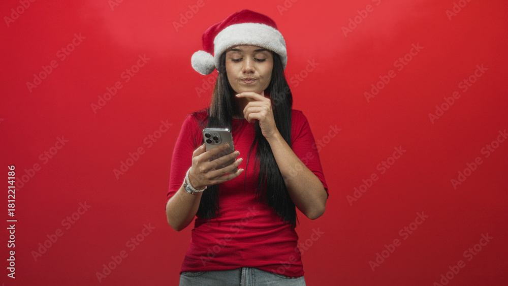 Fototapeta premium Young woman in santa hat holds smartphone, reads screen and touches chin against red backdrop in studio; thoughtful planning.
