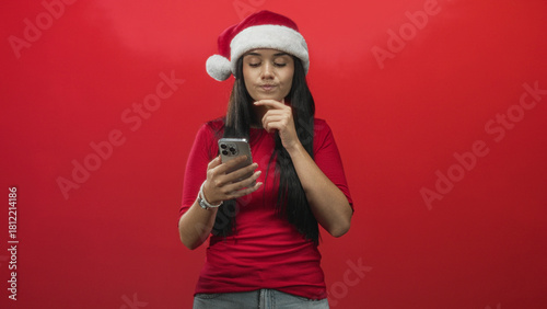 Young woman in santa hat holds smartphone, reads screen and touches chin against red backdrop in studio; thoughtful planning.