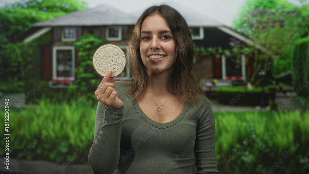 Naklejka premium Woman holding ricecake between fingers at house entrance building, smiling while presenting the round snack to camera; healthy snack contentment.