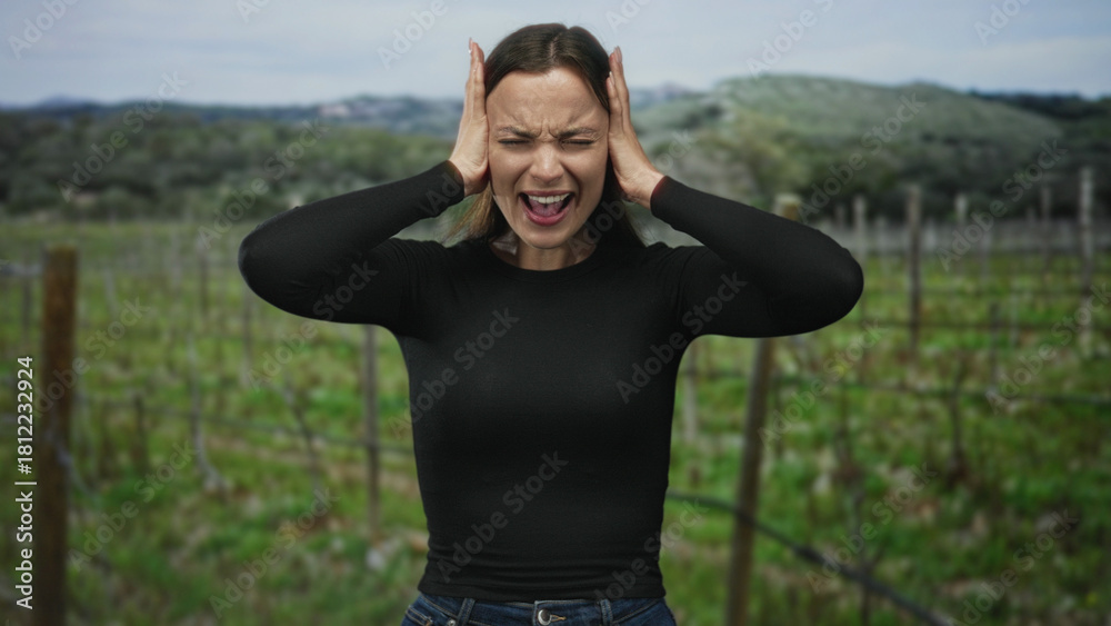 Obraz premium Woman in black top and jeans pressing hands to ears, eyes closed and mouth open in forest vineyard; emotional distress.