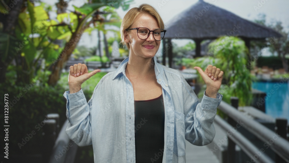 Fototapeta premium Woman pointing thumbs at herself on resort bridge building wearing glasses and casual shirt, smiling broadly with relaxed pose; confidence joy.