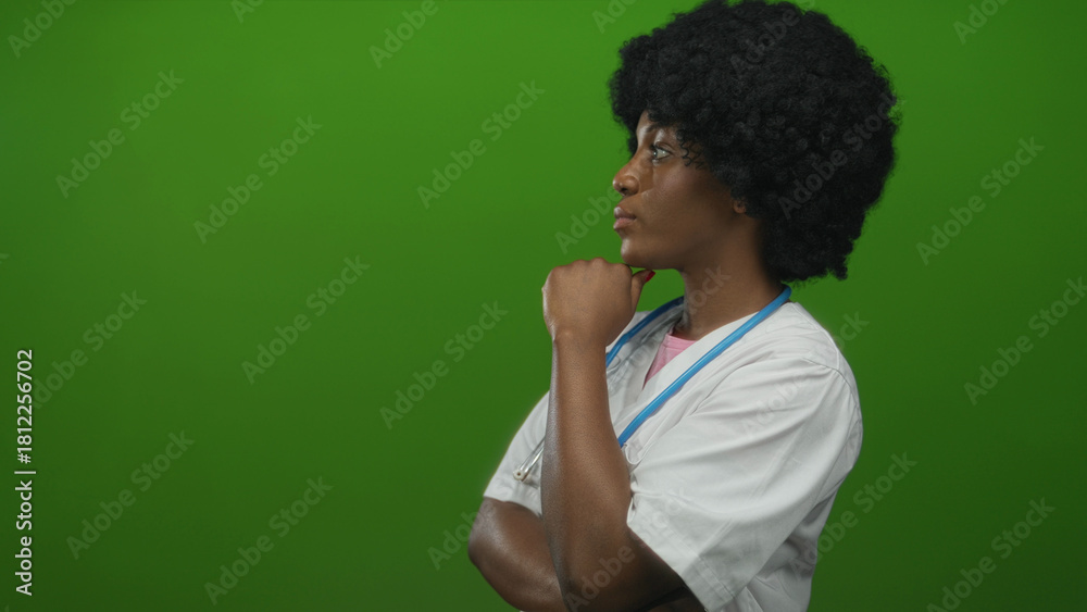 Fototapeta premium African american woman doctor holds stethoscope while hand on chin in green studio; serious contemplation.