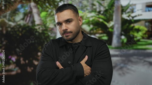 Hispanic man wearing a black jacket with serious expression pointing finger forward in a green park; warning.