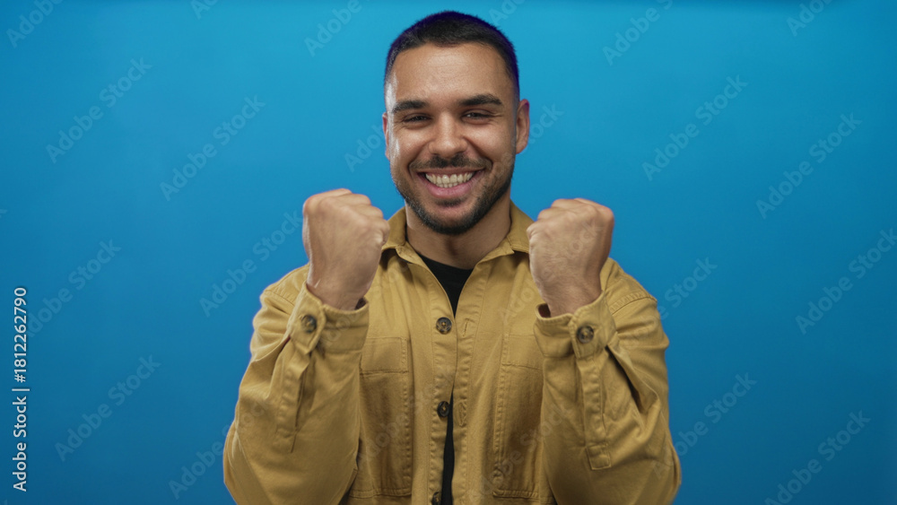 Fototapeta premium Man grins and clenches fists with broad smile in vibrant blue studio setting; victory celebration energy passion.