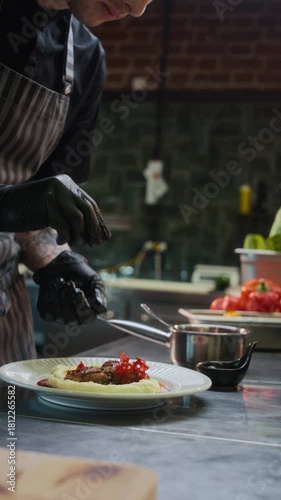 Vertical slow motion shot of young professional chef sprinkling garnish on top of dish while plating food in restaurant kitchen