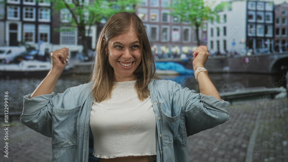 Fototapeta premium Young blonde woman raises clenched fists in front of canal water and brick buildings on street in amsterdam; victory joy.