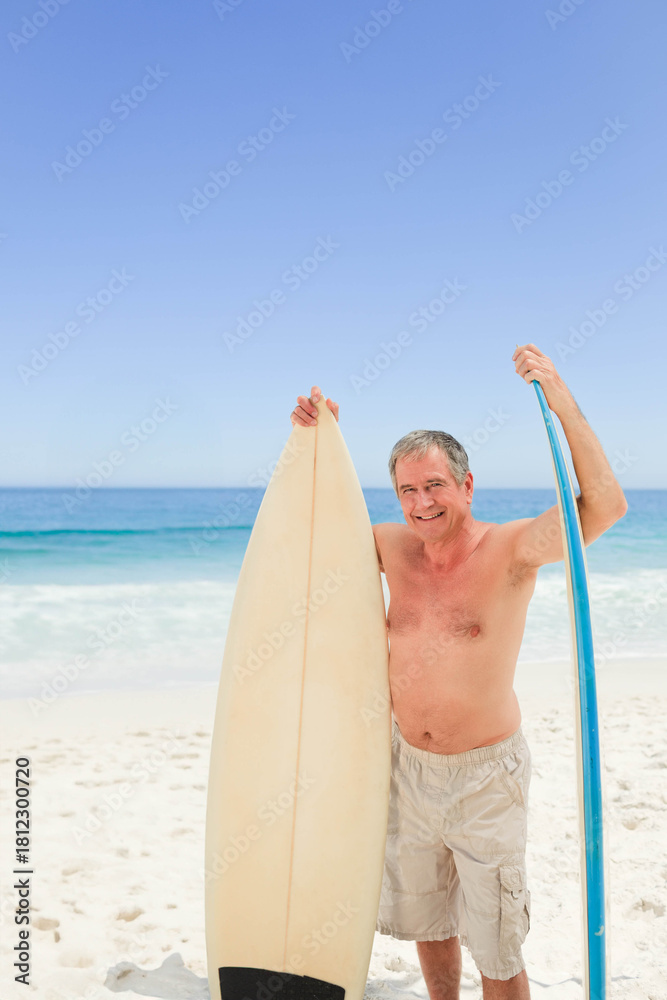 Fototapeta premium Senior man shirtless standing on sandy beach holding off-white and blue surfboards by calm ocean