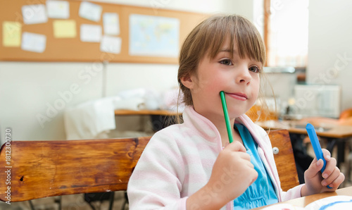Female child sitting at school wooden bench desk wearing pink jacket holding green and blue markers