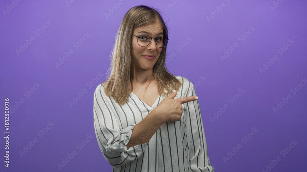 Fototapeta premium Young blonde woman wearing round glasses and striped shirt pointing finger to the right in purple studio; hush silence calm stillness.