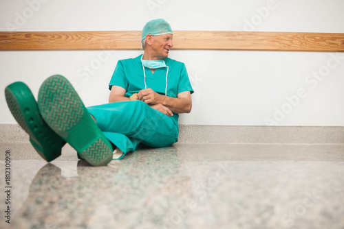 Fotografie Senior male surgeon sitting in hospital hall leaning on wall by rail wearing scr
