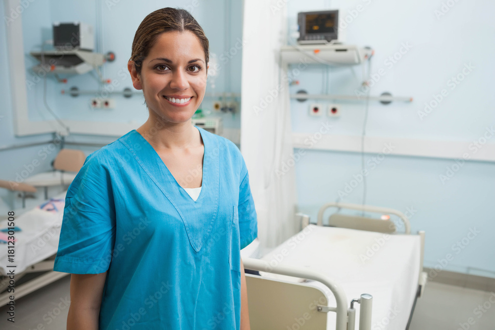 Fototapeta premium Female nurse in blue scrubs standing and smiling, checking monitor and bed rails in hospital room