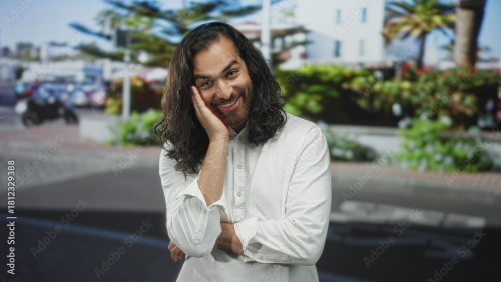 Fototapeta premium Man with long hair and beard smiling, hand to cheek and folded arm on city street near planter and parked scooter; shy joy.