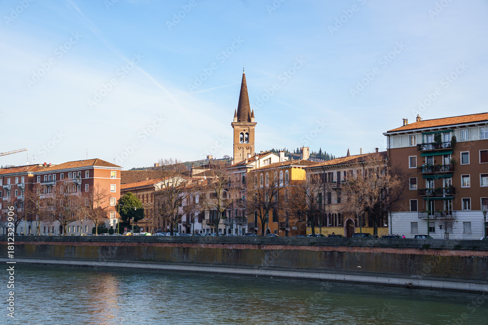 Fototapeta premium A view of part of the city on the waterfront in Verona, Italy. The tall, pointed bell tower of the church is visible