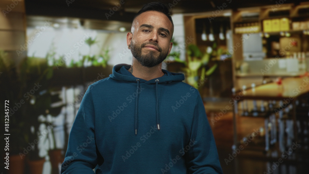 Fototapeta premium Man with exposed face and beard wearing teal hoodie standing with hands clasped in a restaurant building; confidence casual attitude.