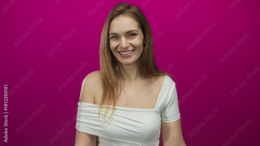 Obraz premium Young woman smiling with bare shoulder and pointing finger to temple in a studio with pink background; confidence.